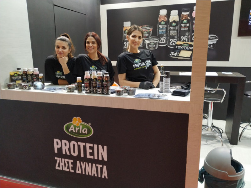 τρεις κοπέλες promoter της Concept on Action στέκονται πίσω από stand της Arla και κάνουν γευστική δοκιμή στα προϊόντα Arla Protein στους επισκέπτες του Ημιμαραθώνιου Αθηνών 2019 στην HELEXPO